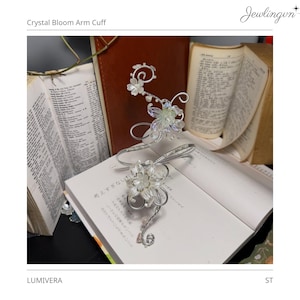 Puede incluir: Un brazalete plateado con un diseño floral, con cristales transparentes y delicados pétalos. El brazalete está adornado con un grupo de flores y descansa sobre un libro abierto. Incluye el texto "Crystal Bloom Arm Cuff".