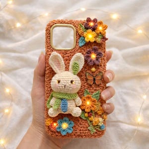 Puede incluir: Funda de teléfono de ganchillo naranja adornada con un conejito 3D con bufanda verde y corbata azul. La funda está decorada con flores de ganchillo coloridas y una mariposa, creando un diseño caprichoso y artesanal.