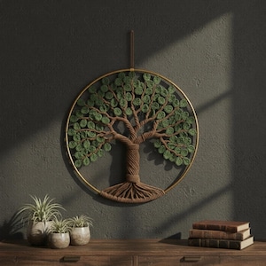 Puede incluir: Un colgante de pared hecho a mano con un diseño de árbol. El árbol tiene un tronco marrón y hojas verdes, encerrado en un marco circular de color dorado. La obra de arte está suspendida contra una pared gris oscuro, con una superficie de madera debajo.