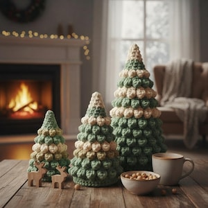 Può includere: Tre alberi di Natale fatti a mano all'uncinetto, di varie dimensioni, con filato verde e crema. Due piccole statuette di renne in legno sono davanti agli alberi. Una piccola ciotola di noci e una tazza di caffè sono sul tavolo di legno.