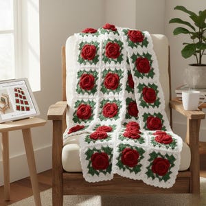 May include: A white crocheted blanket with red rose appliqués and green leaves is draped over a wooden armchair. The blanket is made of square blocks. A tablet displaying a similar blanket is on a side table.