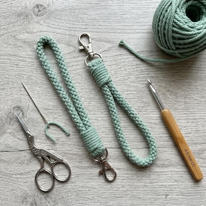 May include: A close-up shot of crafting supplies, including two mint green lanyards with silver clasps, a ball of yarn, a crochet hook, scissors, and a needle. The hook is 6.0 mm (J).