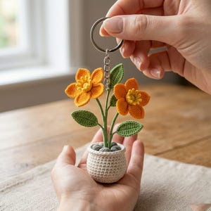 May include: A handmade crochet keychain featuring a small potted plant with two orange and yellow flowers, green leaves, and a beige pot. The keychain is held by a silver ring and chain.
