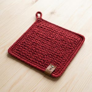 May include: A square, red crocheted potholder with a loop for hanging. The potholder has a textured surface and a small leather tag with text. The potholder is approximately 8 inches square.