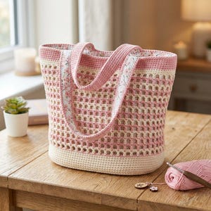 May include: A handmade crochet tote bag in a pink and cream color scheme. The bag features a lattice design, with pink straps adorned with a floral pattern. A ball of pink yarn and a crochet hook are next to the bag.