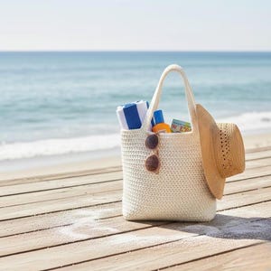 May include: A cream-colored, crocheted tote bag filled with beach essentials. The bag holds a striped towel, sunscreen, sunglasses, and a straw hat. The scene is set on a wooden boardwalk with the ocean in the background.
