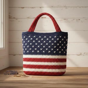 Pode incluir: Uma sacola de crochê com um design patriótico. A sacola apresenta uma parte superior azul marinho com pontos de cruz brancos, listras vermelhas e brancas e uma alça vermelha. A sacola é feita de fio.