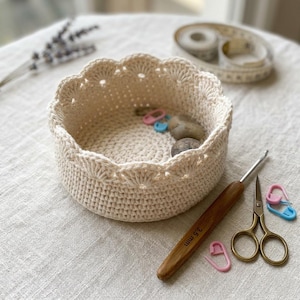 Puede incluir: Una cesta de ganchillo de color crema con borde festoneado, que contiene pequeñas piedras y marcadores de punto. También hay un ganchillo, unas tijeras y una cinta métrica, lo que sugiere un tema artesanal. El ganchillo está marcado con "3,5 mm".