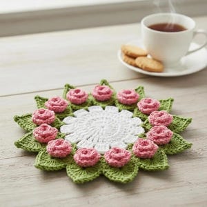 May include: A crocheted doily with a floral design. The doily has a white centre, green leaves, and pink roses. A white teacup with tea and biscuits is in the background.