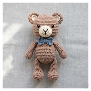 Può includere: Un orsacchiotto all'uncinetto fatto a mano, nei toni del marrone, con muso e orecchie color crema. Presenta un papillon blu e occhi di sicurezza neri. L'orso misura circa 25 cm di altezza.