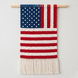 May include: A macrame wall hanging of the American flag. The flag design includes a blue field with white stars, red and white stripes, and a fringe. It is mounted on a natural wooden dowel.