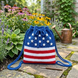 May include: A blue crochet drawstring bag with white stars and red and white stripes. The bag has blue drawstrings and is displayed outdoors with flowers and a watering can in the background.
