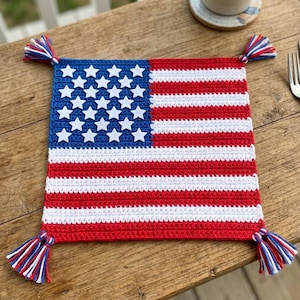 May include: A crocheted square featuring the American flag design. The blue field has white stars, and the rest of the square has red and white stripes. Each corner has a tassel with red, white, and blue threads. The square measures approximately 10 inches by 10 inches.