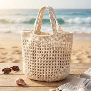 Op de afbeelding: Een crèmekleurige, open geweven gehaakte tote bag met twee handvatten. De tas staat op een houten oppervlak naast een zonnebril en een zeeschelp. De achtergrond toont een strand en de oceaan.