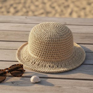 Op de afbeelding: Een beige, gehaakte zonnehoed met een ronde kroon en een brede rand. De hoed is gemaakt van een geweven materiaal en ligt op een houten oppervlak naast een zonnebril met schildpadmotief en een kleine zeeschelp.