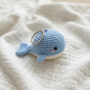 May include: A light blue crocheted whale keychain with a silver keyring. The whale has a white belly, small fins, and a tail. The whale's eyes are black, and it has a small pink mouth. The keychain is lying on a white, textured surface.