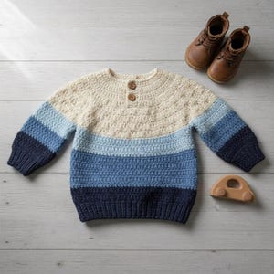 Może przedstawiać: Ręcznie robiony sweter z szydełka w odcieniach kremu, jasnoniebieskiego i granatowego. Sweter ma okrągły dekolt z dwoma drewnianymi guzikami i prążkowanymi mankietami. Na zdjęciu znajdują się również brązowe skórzane buciki dla dziecka i drewniany samochodzik.