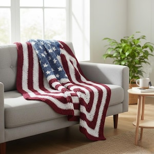 Könnte beinhalten: Eine gehäkelte Decke, die über einem grauen Sofa drapiert ist und ein Design aufweist, das an die amerikanische Flagge erinnert. Die Decke zeigt rote und weiße Streifen mit einem blauen Quadrat, das mit weißen Sternen verziert ist. Die Decke ist aus weichem Garn.