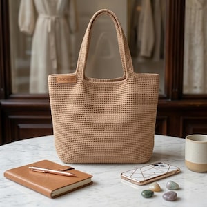 May include: A beige crocheted tote bag with a rectangular shape and two handles. The bag has a label that says "CROCHETED". A brown leather notebook with a pen, a smartphone, and a ceramic mug are on the table.