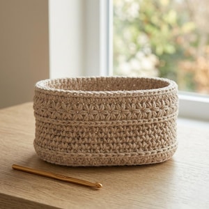 Könnte beinhalten: Ein beige, handgehäkelter Korb mit einem dekorativen Blumenmuster in der Mitte. Der Korb ist oval geformt und steht auf einer Holzoberfläche. Ein goldener Häkelhaken liegt neben dem Korb auf dem Tisch.