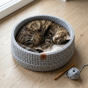 May include: A gray, round, crocheted cat bed with a sleeping tabby cat inside. The bed has a soft, white interior and a brown leather tag. A ball of gray yarn and a wooden crochet hook are next to the bed.