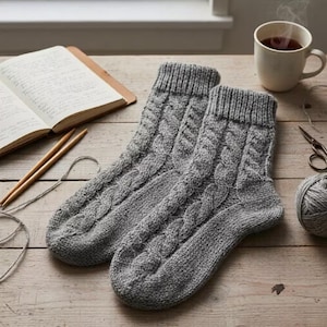 Op de afbeelding: Een paar grijze kabelgebreide sokken, liggend op een houten oppervlak. De sokken hebben een textuurontwerp en liggen naast een notitieboek, breinaalden, een kop thee, een schaar en een bol garen. De scène suggereert een gezellig, handgemaakt ambacht.