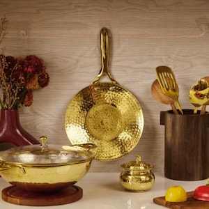 Puede incluir: Una colección de utensilios de cocina dorados, que incluyen una sartén, un cuenco y un tarro pequeño, dispuestos sobre una superficie blanca. También hay un jarrón rojo oscuro con flores secas y un soporte de madera para utensilios. Una tabla de cortar de madera con verduras está en primer plano.