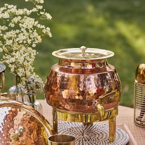 May include: A hammered copper beverage dispenser with a brass lid, spigot, and stand. The dispenser sits on a woven mat, with a blurred background of greenery and flowers. The dispenser is likely used for serving drinks at a party or event.