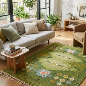 May include: A living room scene featuring a light gray sofa with decorative pillows, a wooden side table, and a green patterned rug. The rug has a fringe and floral accents. Large windows and potted plants add to the room's decor.