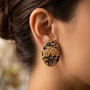 May include: An oval-shaped earring featuring a black background with intricate, raised gold-toned floral detailing. The design displays multiple blooming flowers and leaves in a relief pattern. The piece is secured to the earlobe with a post backing.