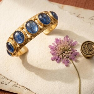 Pu&ograve; includere: Un bracciale rigido dorato con cinque pietre preziose di cianite blu di forma ovale incastonate singolarmente. Il bracciale presenta una finitura metallica testurizzata ed &egrave; accompagnato da un sigillo rotondo in metallo e un fiore viola essiccato.
