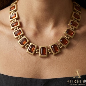 May include: A necklace featuring a series of rectangular, matte gold-toned metal links, each set with a faceted, amber-colored rectangular gemstone. The links are connected by small, double-bar hinges, creating a structured, articulated collar design.