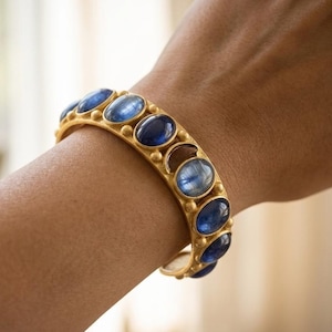 Pu&ograve; includere: Un bracciale rigido color oro caratterizzato da una serie di pietre preziose di cianite blu intenso di forma ovale. La fascia in metallo ha una finitura opaca ed &egrave; accentuata da piccole sfere dorate in rilievo tra ogni pietra blu lucida.