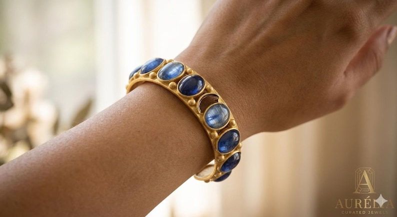 Pu&ograve; includere: Un bracciale rigido color oro caratterizzato da una serie di pietre preziose di cianite blu intenso di forma ovale. La fascia in metallo ha una finitura opaca ed &egrave; accentuata da piccole sfere dorate in rilievo tra ogni pietra blu lucida.