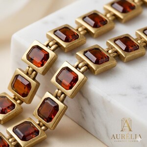May include: A bracelet featuring a series of rectangular, matte gold-toned metal links, each set with a deep amber-colored, faceted gemstone. The piece includes the text AUR&Eacute;LIA CURATED JEWELS in the bottom right corner of the frame.
