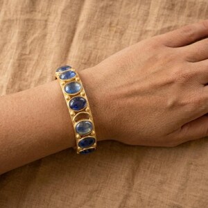 Pu&ograve; includere: Un bracciale rigido color oro caratterizzato da una serie di pietre blu lucide di forma ovale, taglio cabochon, disposte in fila. La fascia metallica ha una finitura opaca e mostra un motivo ripetitivo di castoni circolari per ogni gemma.