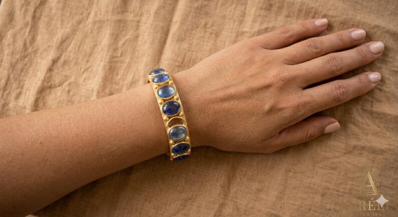 Pu&ograve; includere: Un bracciale rigido color oro caratterizzato da una serie di pietre blu lucide di forma ovale, taglio cabochon, disposte in fila. La fascia metallica ha una finitura opaca e mostra un motivo ripetitivo di castoni circolari per ogni gemma.