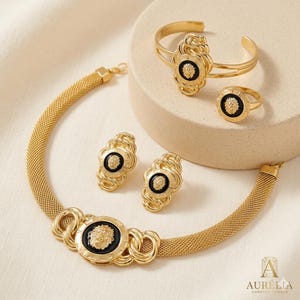 May include: A gold-toned jewelry set featuring a necklace, earrings, ring, and bracelet. Each piece has a lion's head emblem on a black background. The necklace has a mesh chain design, and the bracelet is a cuff style.