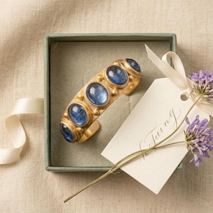 Pu&ograve; includere: Un bracciale rigido color oro con cinque pietre blu ovali a cabochon, presentato in una scatola quadrata verde salvia. Un cartellino regalo color crema con testo in corsivo &egrave; legato alla scatola con un nastro bianco, insieme a due fiori viola.