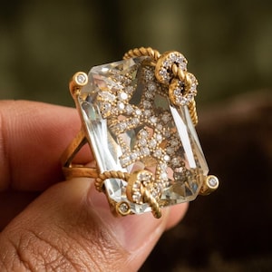 May include: A gold ring featuring a large, rectangular, clear faceted gemstone. Inside the stone is a textured, branch-like structure encrusted with small white stones. The gold band has a twisted rope design that wraps around the gemstone.