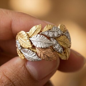 May include: A wide band ring featuring a textured leaf motif in yellow, white, and rose gold tones. The ring is accented with small, round, clear stones embedded within the metallic leaf design, creating a layered and intricate botanical appearance.