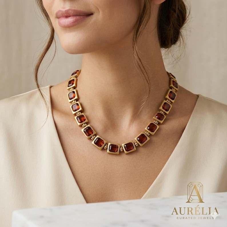 May include: A gold-toned necklace featuring a series of square-shaped, amber-colored glass stones set in matte metal frames. The links are connected by small, round gold beads, creating a uniform chain that rests around the neck area.
