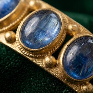 Pu&ograve; includere: Un anello color oro con una serie di pietre cabochon blu intenso di forma ovale incastonate in metallo. La fascia presenta una finitura opaca e testurizzata con piccoli accenti sferici tra ogni incastonatura e un logo in basso a destra.