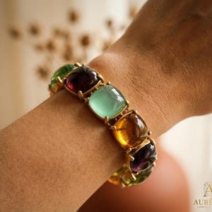 May include: A gold-toned bracelet featuring a series of smooth, square-shaped cabochon stones in various translucent colors including deep purple, light green, and amber. The stones are set in a linked metal frame that wraps around the wrist.