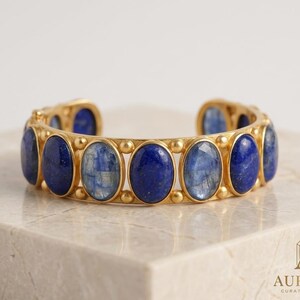Pu&ograve; includere: Un bracciale a fascia color oro caratterizzato da una serie di pietre preziose ovali alternate di lapislazzuli blu e cianite blu, incastonate in una montatura in metallo con piccoli accenti sferici tra ogni pietra. Il logo AUR&Eacute;LIA CURATED JEWELS &egrave; visibile.