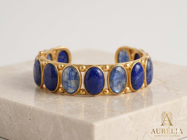 Pu&ograve; includere: Un bracciale a fascia color oro caratterizzato da una serie di pietre preziose ovali alternate di lapislazzuli blu e cianite blu, incastonate in una montatura in metallo con piccoli accenti sferici tra ogni pietra. Il logo AUR&Eacute;LIA CURATED JEWELS &egrave; visibile.
