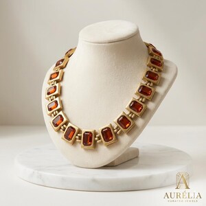 May include: A gold-toned necklace featuring a series of rectangular links, each set with a deep amber-colored rectangular gemstone. The metal has a matte finish, and the links are connected by small hinges, creating a structured, uniform design.
