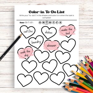 May include: A white "Color-in To-Do List" with heart-shaped boxes for tasks like "call Doc" and "make the bed." A black pencil and a collection of colored pencils are on the wooden surface.