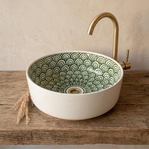 Könnte beinhalten: Ein runder, weißer Keramikwaschtisch mit einem grünen, fächerförmigen Muster im Inneren. Rechts ist ein goldfarbener Wasserhahn montiert. Das Waschbecken steht auf einer Holzoberfläche.