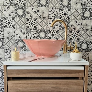 Puede incluir: Un moderno mueble de baño con un lavabo rosa coral con borde festoneado y un grifo dorado. Una pequeña vela y un dispensador decorativo se encuentran sobre la encimera blanca. El fondo presenta azulejos estampados.
